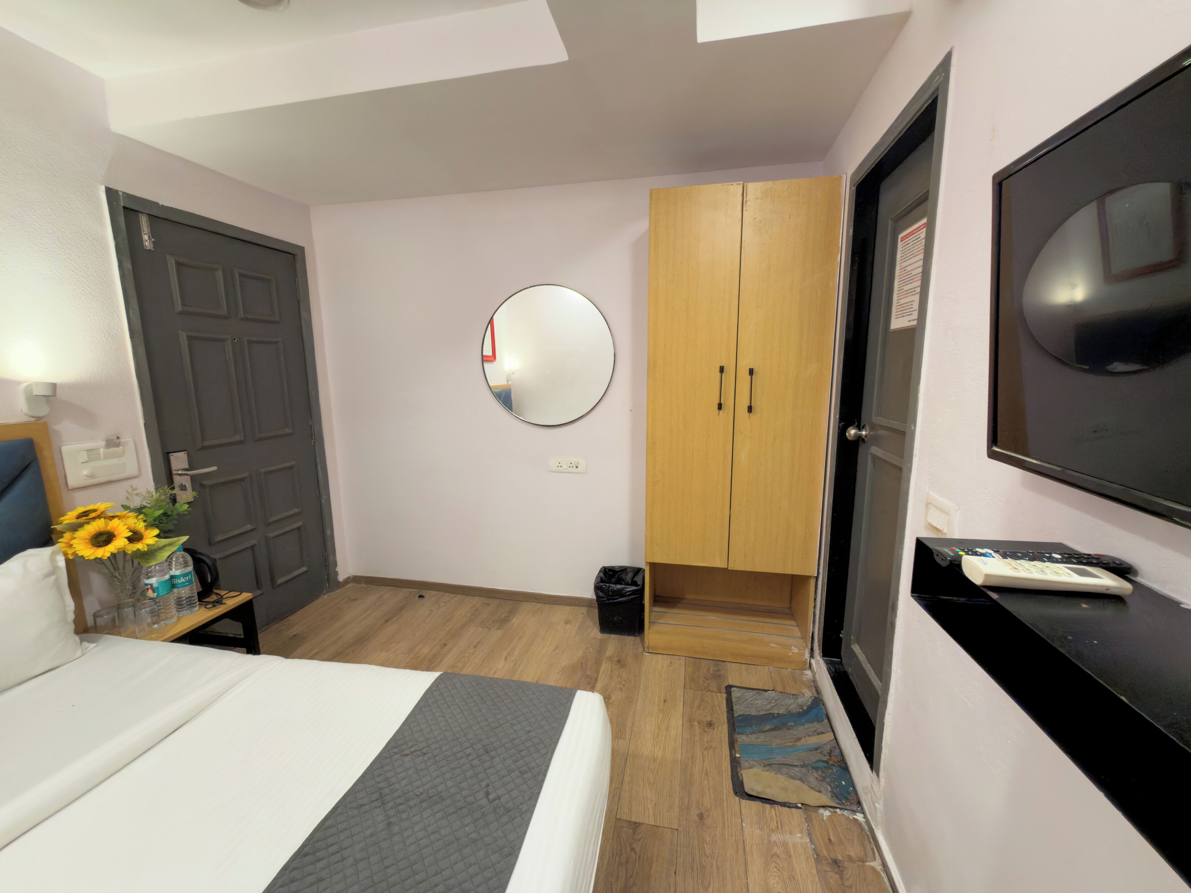 A hotel room with a bed, a TV, a wardrobe, a mirror, and a small table with a vase of sunflowers.