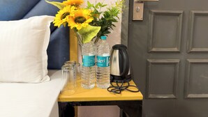 Ein ordentlich bezogenes Bett mit Kissen, ein kleiner Tisch mit einer Vase voller Sonnenblumen, zwei Wasserflaschen und einem Wasserkocher.