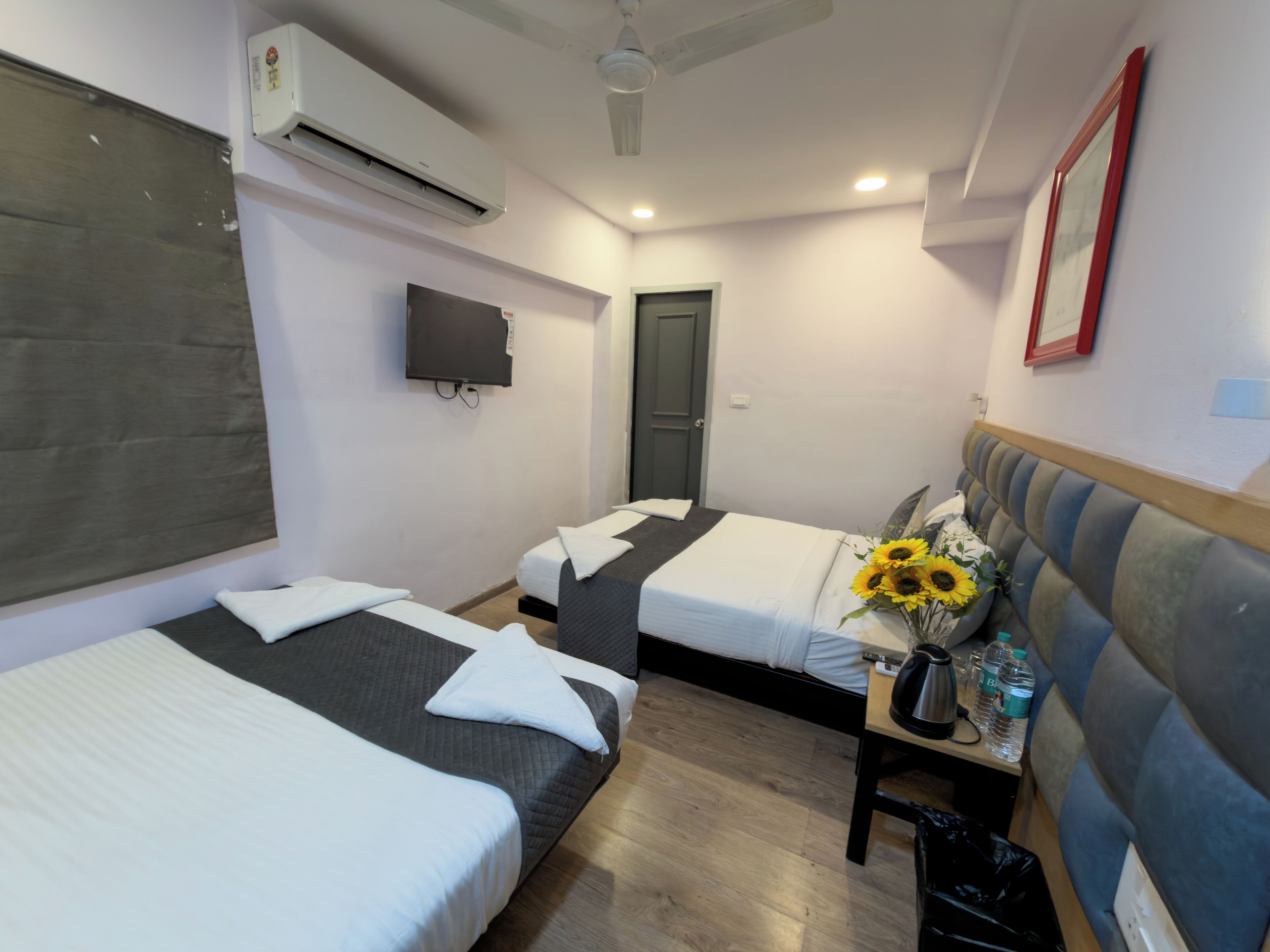A hotel room with two beds, a TV, an air conditioner, a ceiling fan, a door, and a small table with a vase of sunflowers.