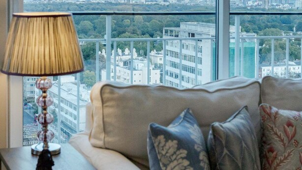 A living room with a sofa, a lamp, and a view of a cityscape through large windows. - AVAL Lovely 2-bed flat with balcony in Notting Hill, West London (London)