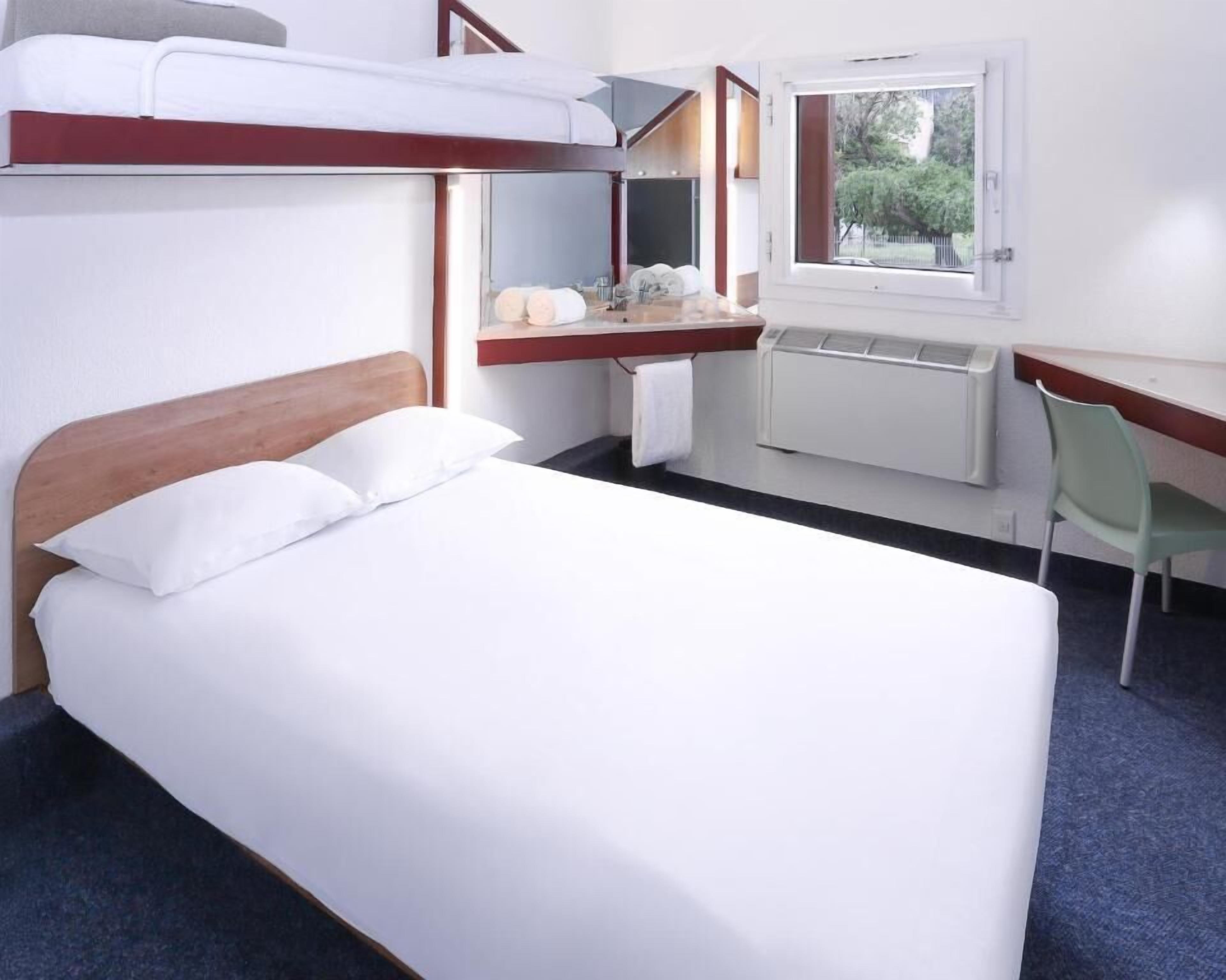 A hotel room with a bunk bed, a desk with a chair, a window, and a radiator.