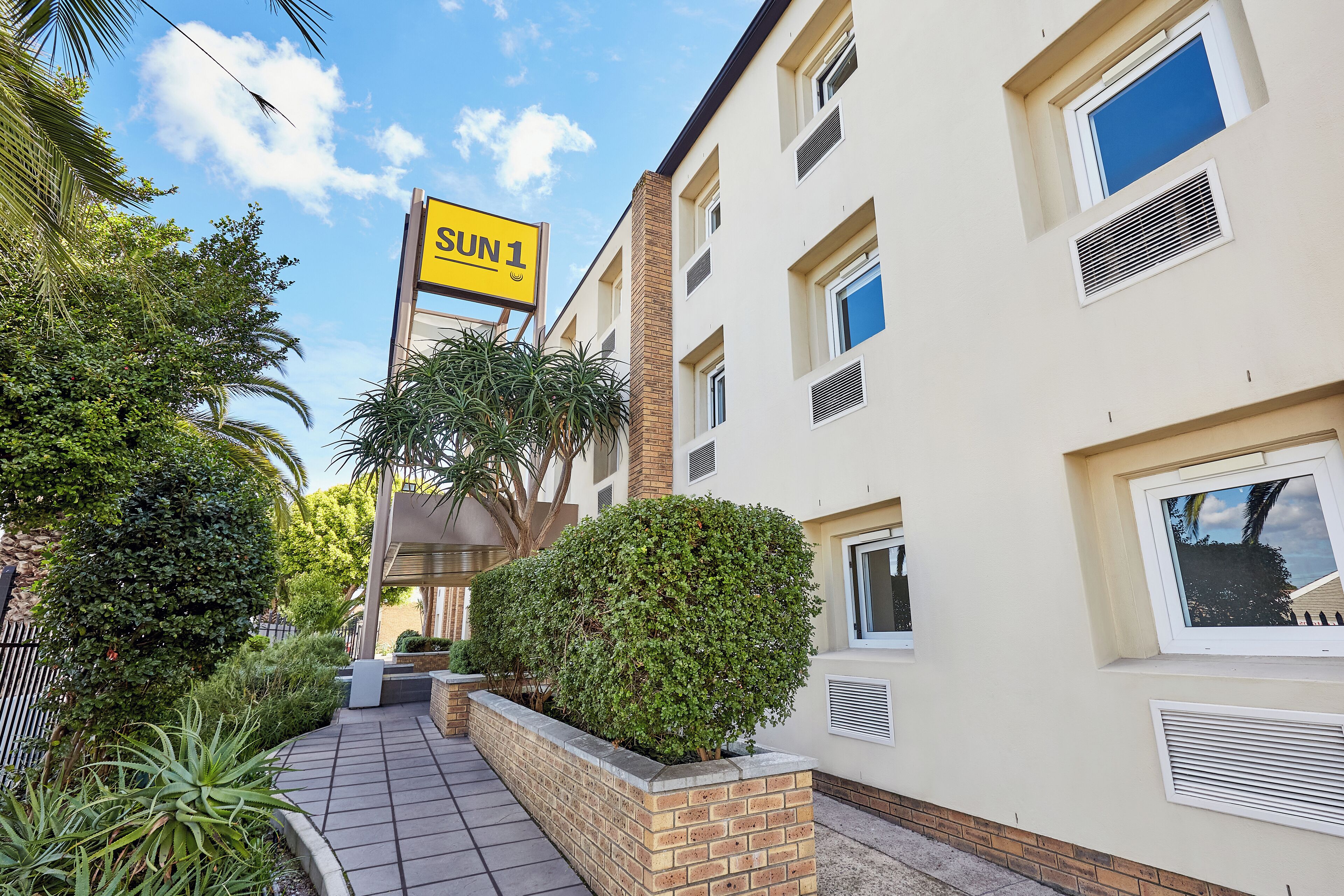A residential building with a 'SUN 1' sign, a brick pathway, and a well-maintained garden area.