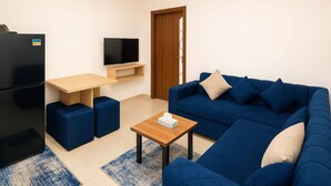Uma sala de estar moderna com um sofá azul, uma geladeira preta, uma mesa de centro de madeira e uma TV em uma prateleira de madeira.