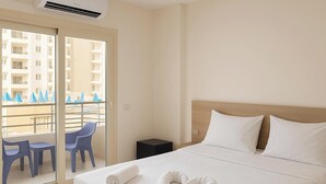 Quarto de hotel com uma cama, uma pequena mesa com cadeiras, uma varanda com vista para edifícios e um ar condicionado.
