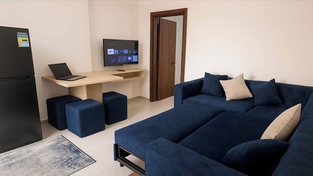 Quarto moderno com sofá azul escuro, uma pequena mesa com laptop e monitor, um frigorífico e uma porta.