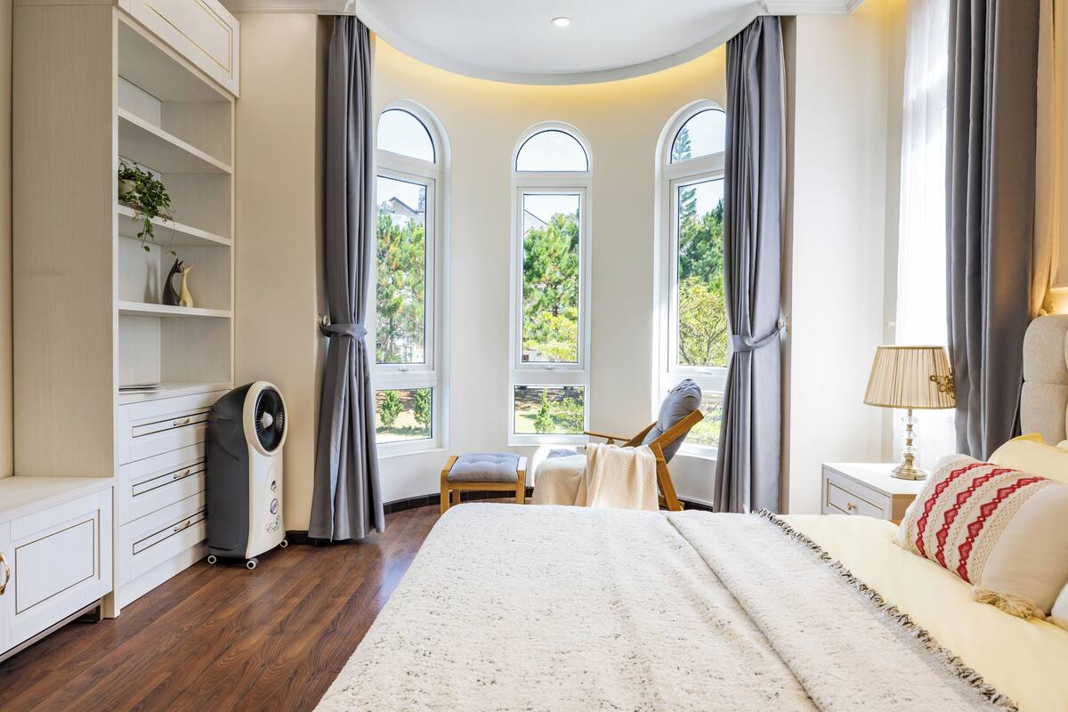 A bedroom with a large bed, a bedside table with a lamp, a chair, a fan, and a window with curtains.