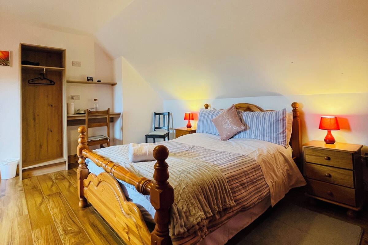 A bedroom with a wooden bed, a nightstand with a lamp, a wooden wardrobe, a desk with a chair, and a small table with a lamp.