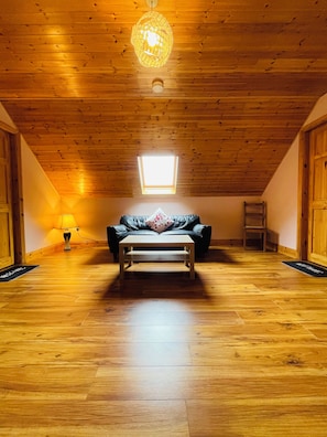 Una habitación con piso de madera, un sofá de cuero negro, una mesita de centro de madera, una lámpara colgante y un tragaluz.