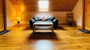 A room with wooden flooring, a black leather sofa, a wooden coffee table, a hanging light fixture, and a skylight. - Tinnies Guest House (Valentia Island)