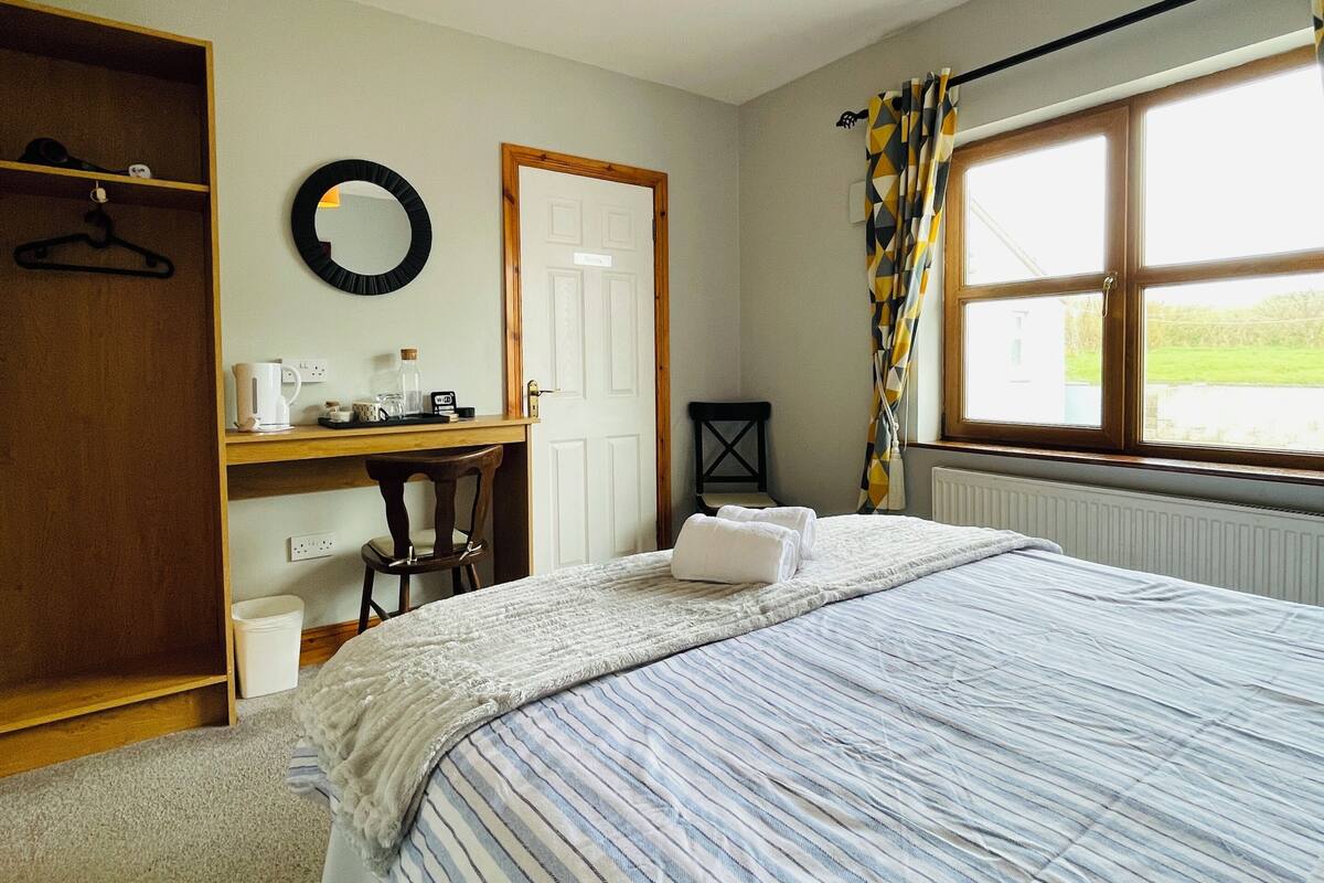 A bedroom with a bed, a desk, a chair, a hanging light fixture, a window with curtains, and a door.