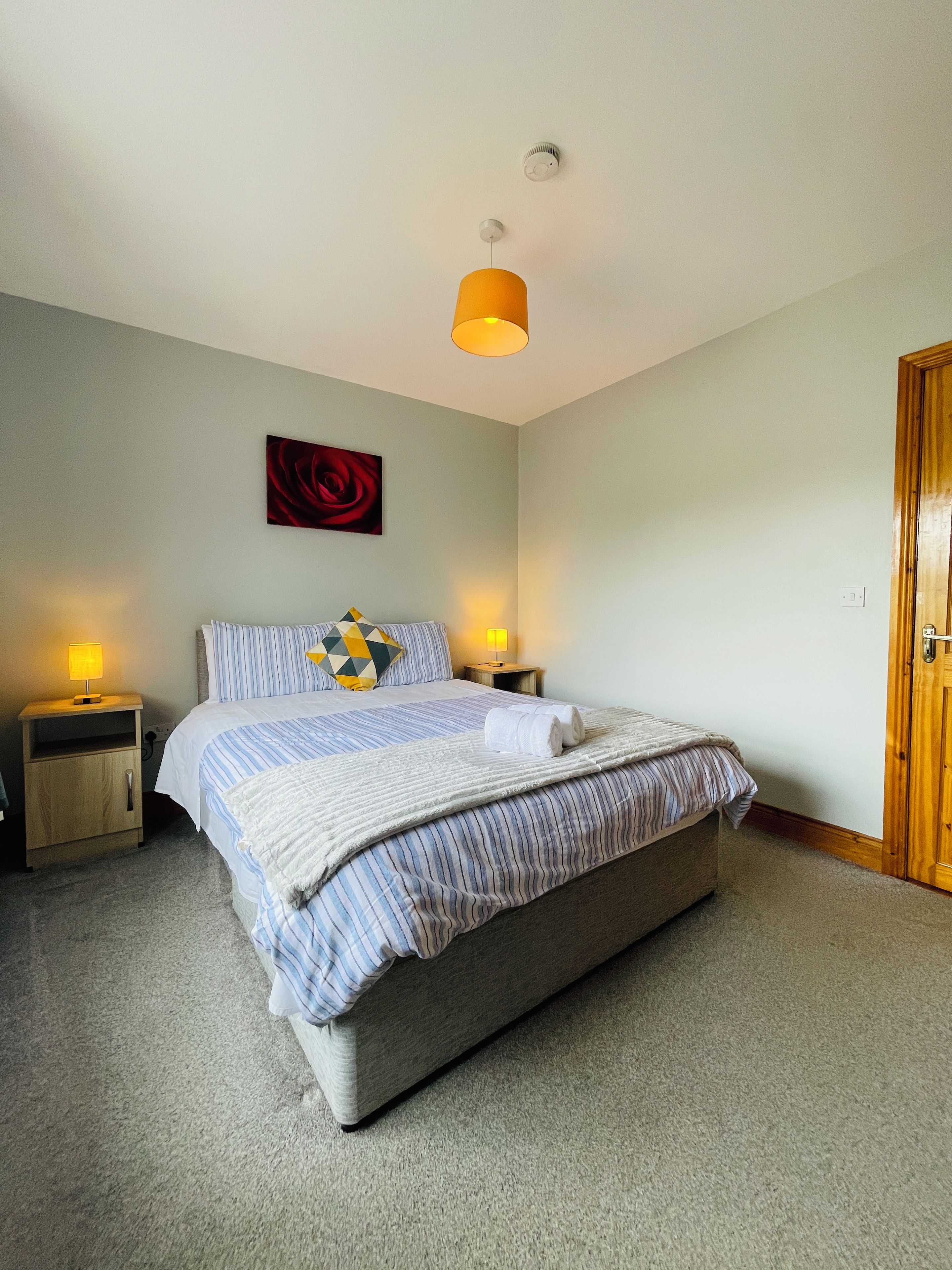 Una cama bien hecha con ropa de cama a rayas y un cojín con un diseño geométrico. Hay dos lámparas de noche de madera. Un cuadro de una sola rosa roja cuelga en la pared sobre la cama.