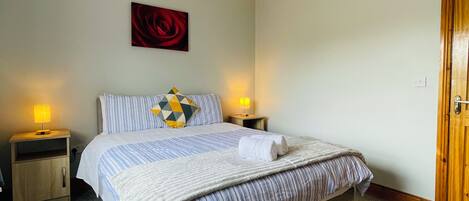 Una cama bien hecha con ropa de cama a rayas y un cojín con un diseño geométrico. Hay dos lámparas de noche de madera. Un cuadro de una sola rosa roja cuelga en la pared sobre la cama.