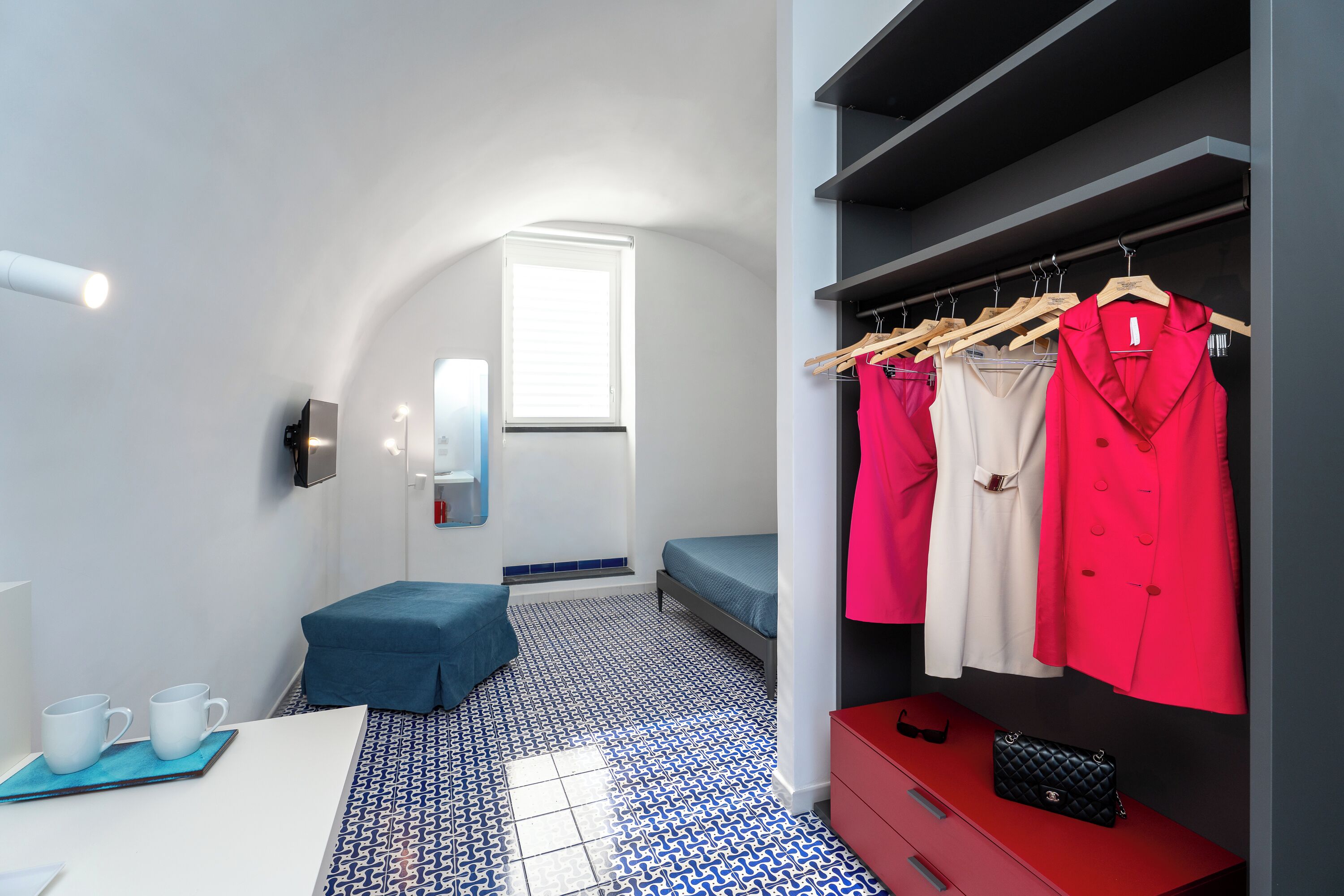 Una habitación de hotel moderna con cama, un banco, un armario con ropa, una mesa con tazas y una ventana.