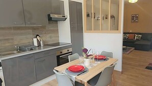 A modern kitchen with grey cabinets, a wooden dining table set for four, and a living area with a sofa and a cabinet. - Large 47 m² renovated family apartment on 1st floor - Portes des Ardennes (Charleville-Mézières)