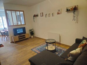 Un salon comprenant un canapé, une table basse, un téléviseur posé sur un meuble, une plante et un mur orné de tableaux.