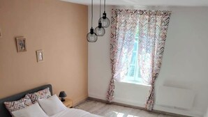 A bedroom with a bed, a nightstand, a lamp, a window with patterned curtains, and a ceiling light fixture. - Large 47 m² renovated family apartment on 1st floor - Portes des Ardennes (Charleville-Mézières)
