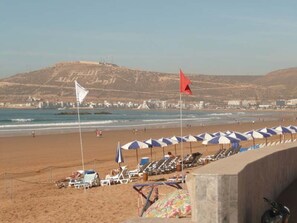 A beach with umbrellas and lounge chairs, a flag, and a hilly background. - Résidence Jet Sakane by Interhome (Agadir)
