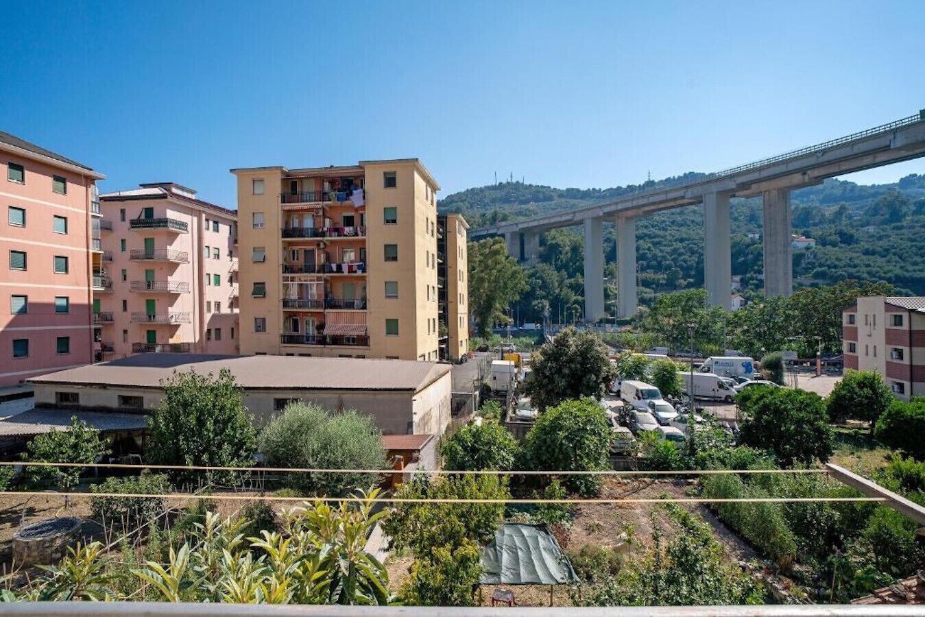 A multi-story residential building with balconies, a parking area with cars, and a bridge in the background.