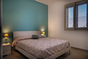 A bedroom with a bed, two bedside lamps, a window with blinds, and teal walls. - Nella's House (Lido di Noto)