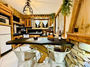 Une cuisine compacte avec des meubles en bois, un plan de travail noir et un mur en pierre. On y trouve un petit coin repas avec des chaises blanches et une table.