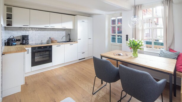Eine moderne Küche mit weißen Schränken, einer Holzarbeitsplatte und einem eingebauten Ofen. Es gibt einen Essbereich mit zwei grauen Stühlen und einem Tisch. Ein Fenster mit Vorhängen ist sichtbar, und auf dem Tisch steht eine Vase mit Blumen.