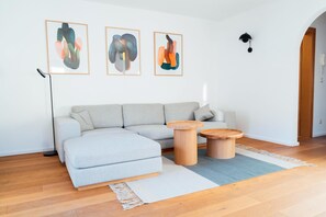 Ein modernes Wohnzimmer mit einem grauen Ecksofa, zwei Holztischen und abstrakter Wandkunst.