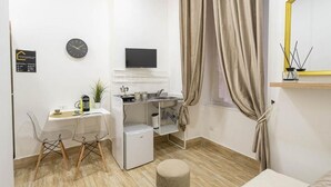 A compact room with a desk, chairs, a small refrigerator, a TV, a clock, and a window with curtains. - Mega Random By Dimorra (Naples)