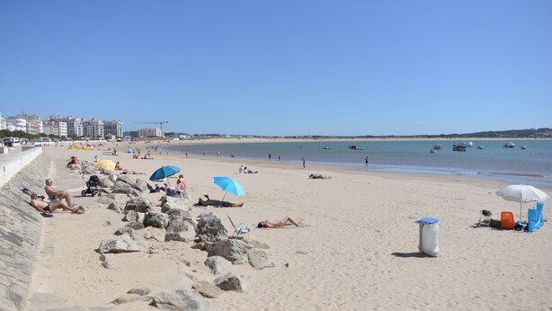 Una playa arenosa con gente tomando el sol y nadando, cielo despejado y embarcaciones en el agua.
