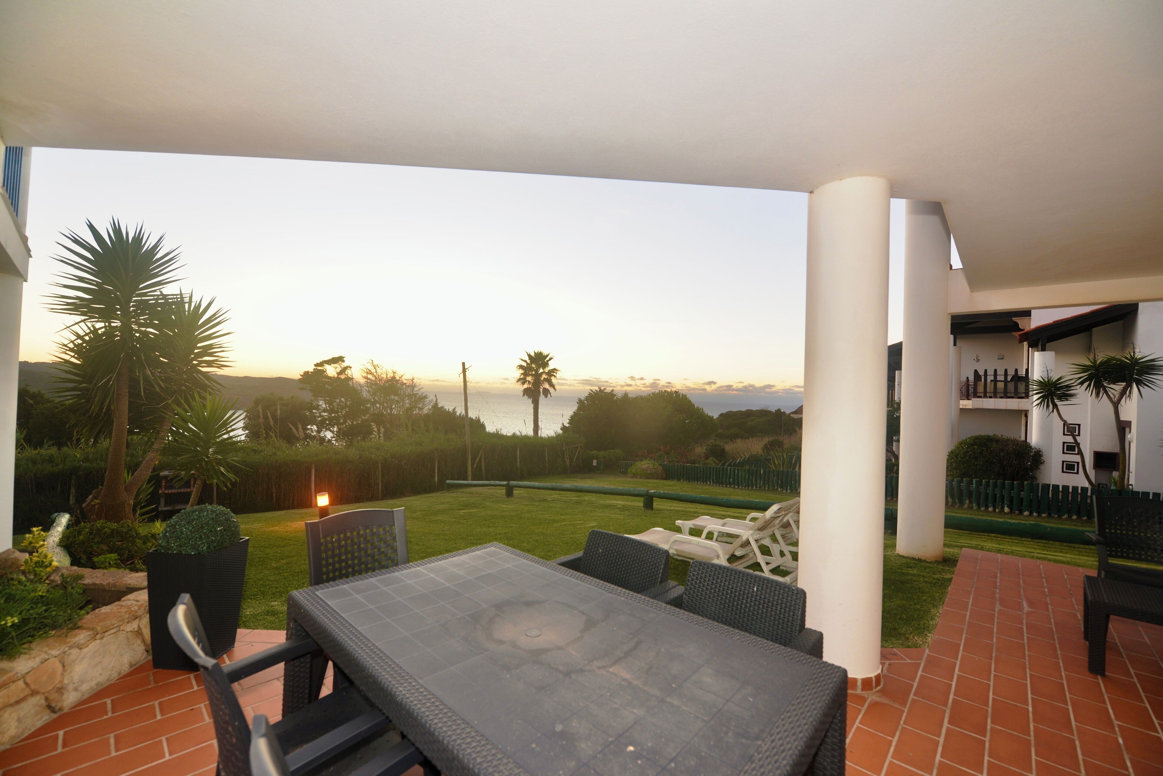 Un espace extérieur aménagé en terrasse, avec une table et des chaises, entouré d’une pelouse bien entretenue et de palmiers.