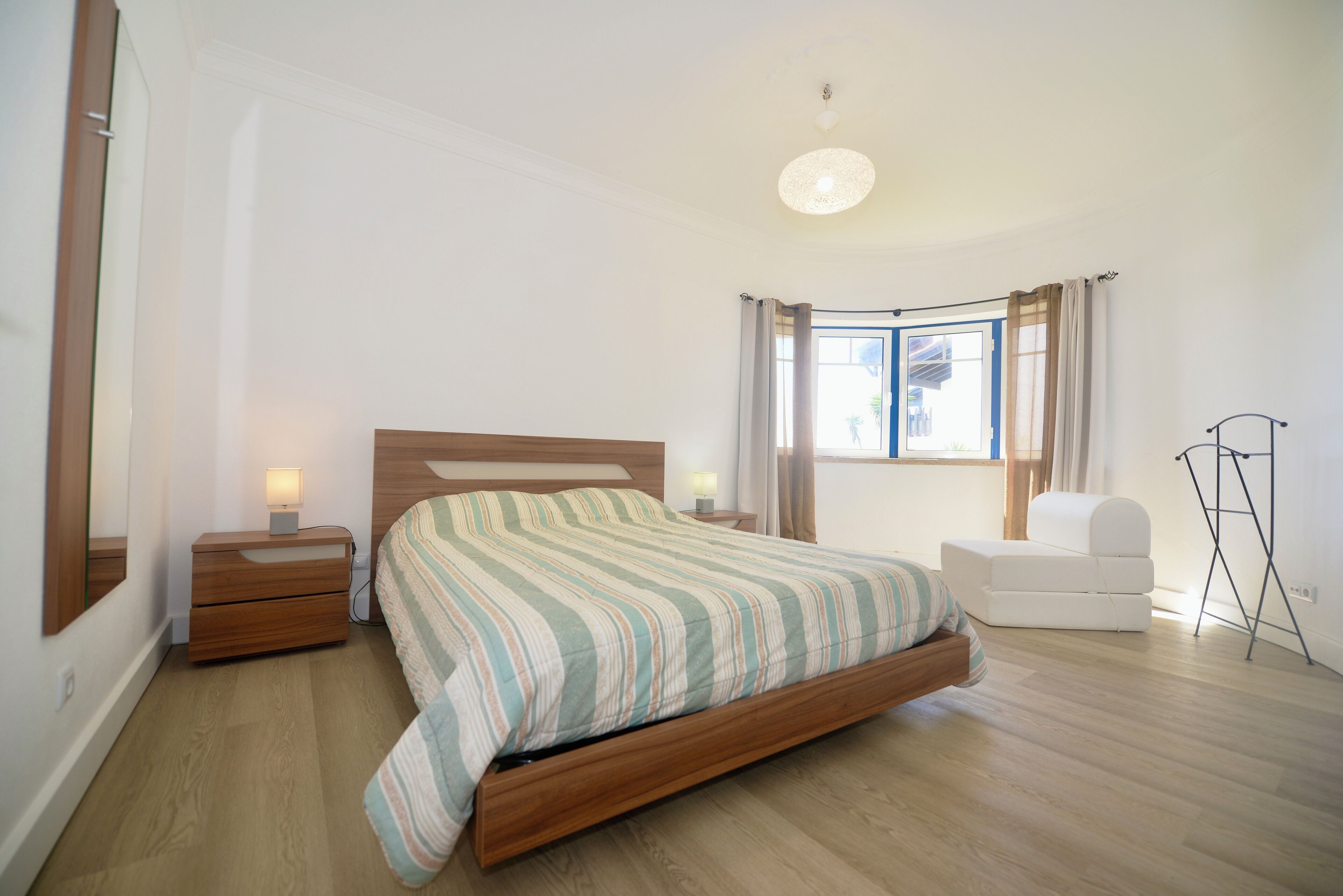 Une chambre à coucher avec un lit en bois, des tables de chevet avec des lampes, une chaise blanche et une fenêtre avec des rideaux.