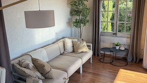 A cozy living room with a sofa, two small tables, a potted plant, and a window with curtains.