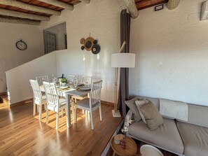 A dining area with a wooden table set for four, surrounded by white chairs. A lamp and wall decorations are visible.