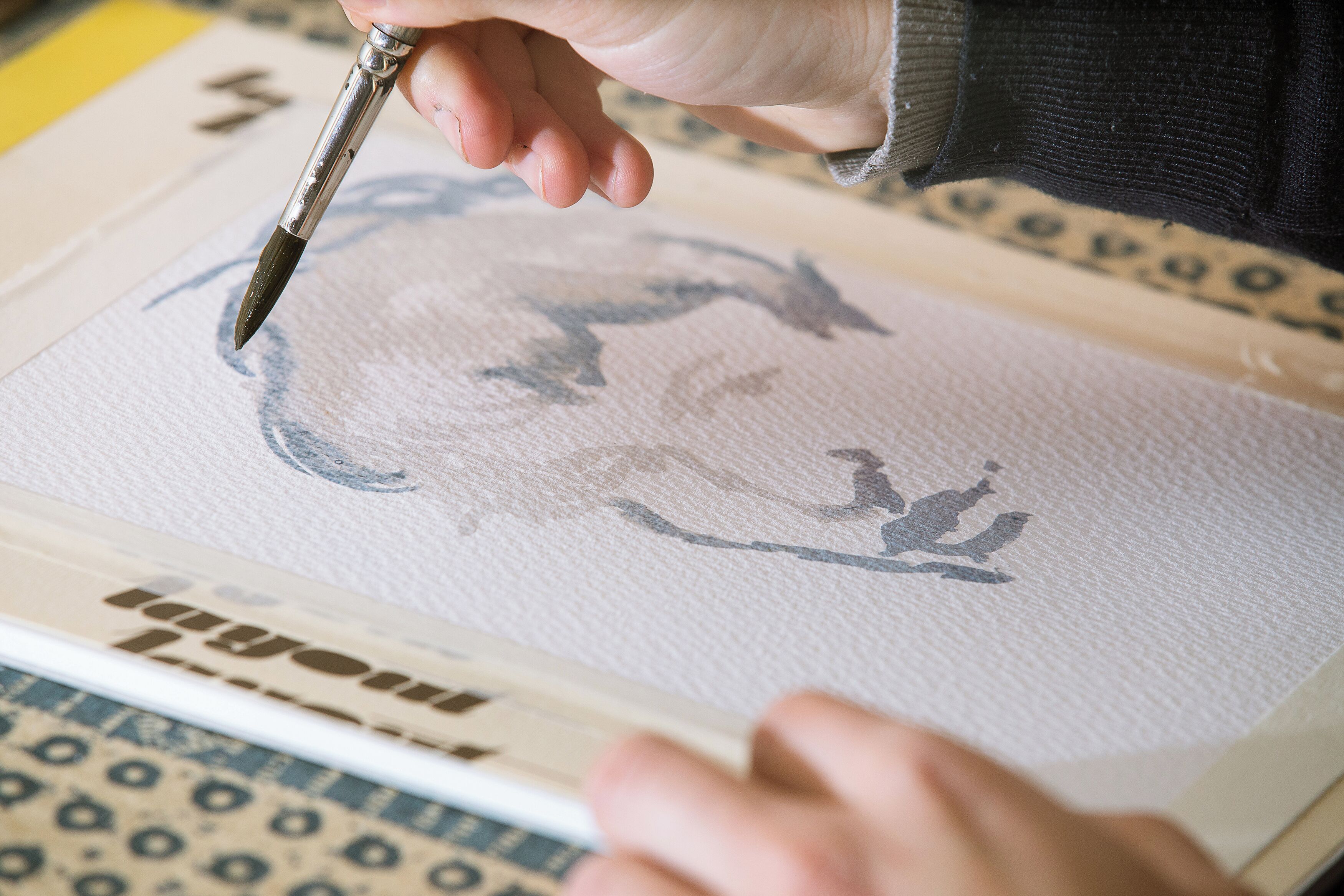 Une main tenant un pinceau, peignant sur un papier texturé orné d’un motif.