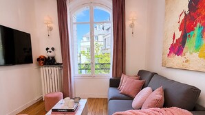 Ein Wohnzimmer mit einer Couch, einem Couchtisch mit Büchern, einem Fernseher, einer Heizung und einem Fenster mit Vorhängen.