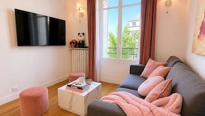 Ein Wohnzimmer mit Flachbildfernseher, einem Sofa mit rosafarbenen und grauen Kissen, einem Couchtisch mit Büchern, einem rosafarbenen Hocker und einem Fenster mit Vorhängen.