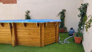 Eine Holzkonstruktion mit blauer Plane als Dach, umgeben von Kunstgrashalme und Topfpflanzen.