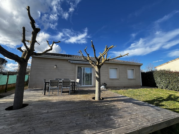 Une maison avec un patio, un coin repas avec des chaises et un arbre au premier plan.