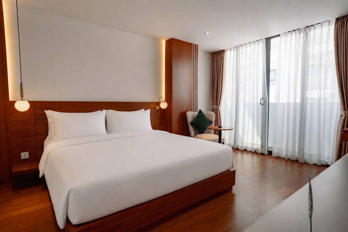 A hotel room with a large bed, bedside lamps, a chair, a small table, and a view of the outdoors through sheer curtains.
