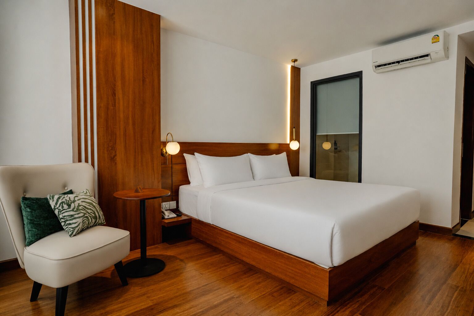 Ein Hotelzimmer mit einem hölzernen Kopfteil, einem Einzelbett, einem Nachttisch mit Lampe, einem Sessel mit Kissen und einer Klimaanlage.
