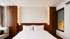 A hotel room with a large bed, wooden headboard, two bedside lamps, a nightstand, a window, and a wall-mounted air conditioning unit. - The Scenic Hotel  (Phnom Penh)