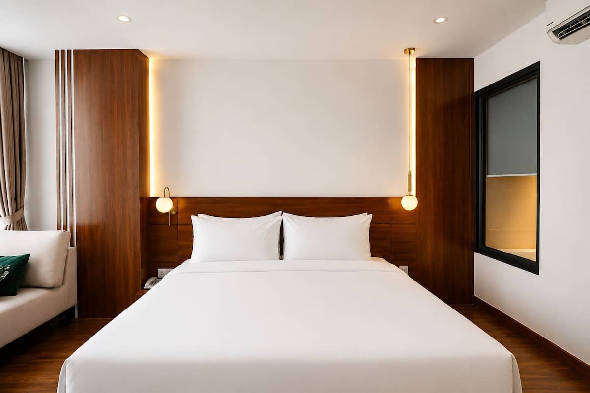 A hotel room with a large bed, wooden headboard, two bedside lamps, a nightstand, a window, and a wall-mounted air conditioning unit.