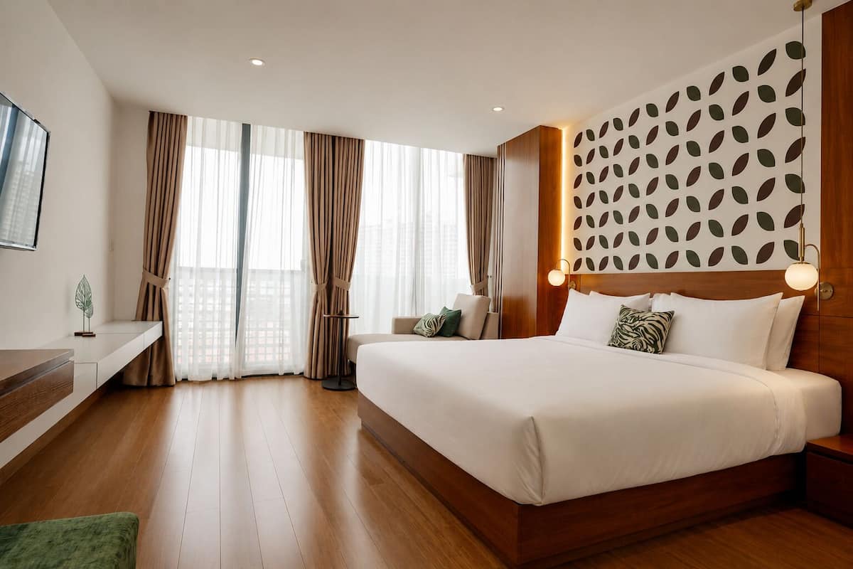A modern hotel room with a large bed, a bedside table, a small table, and a view of the city through large windows.
