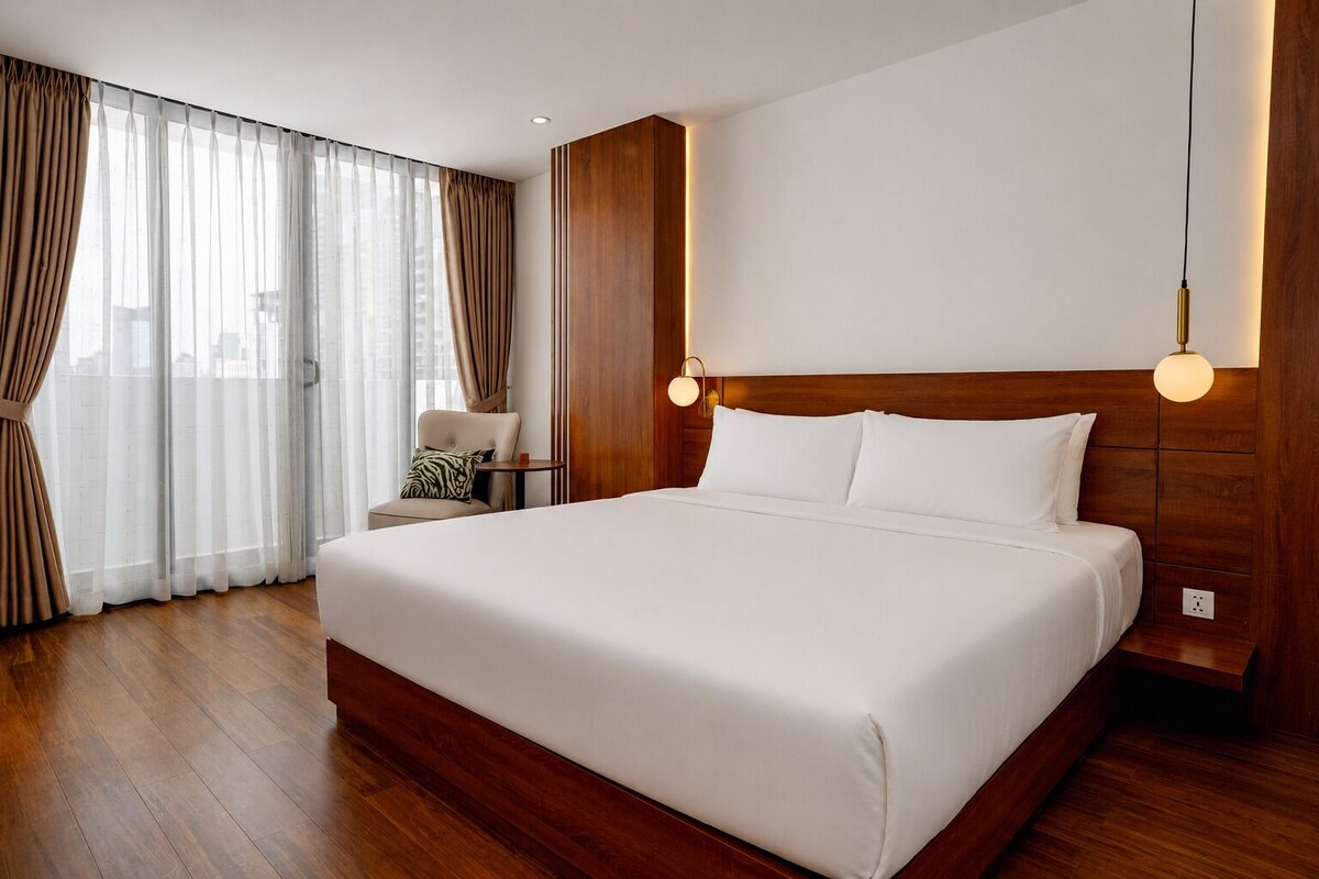 A hotel room with a large bed, wooden headboard, a chair, a small table, and a view of the city through sheer curtains.