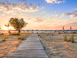 A wooden boardwalk leading to a beach with umbrellas and chairs, a tree, and a sunset in the background. - Georgioupolis Blaue Villa Privater Pool by Interhome (Georgioupoli)