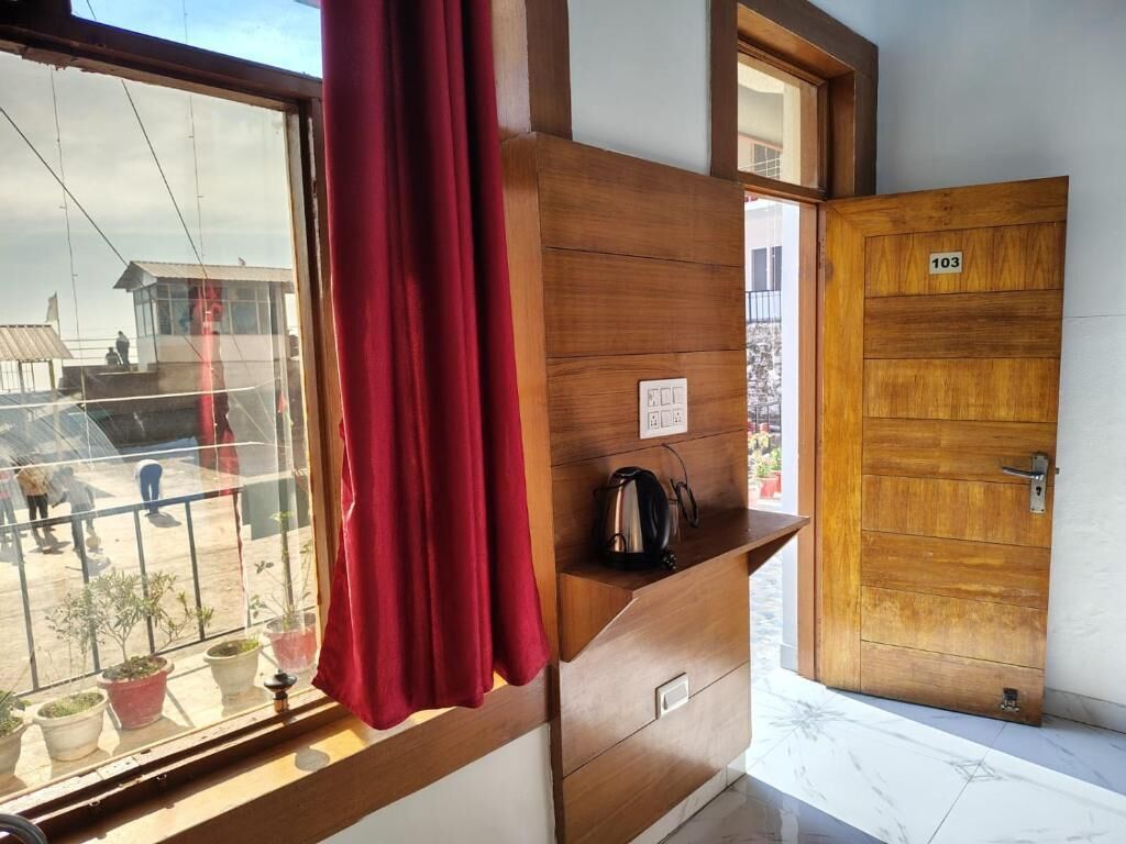 Quarto com porta de madeira identificada com o número '103', janela com cortinas vermelhas e uma pequena mesa com uma chaleira.