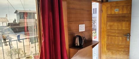 Quarto com porta de madeira identificada com o número '103', janela com cortinas vermelhas e uma pequena mesa com uma chaleira.