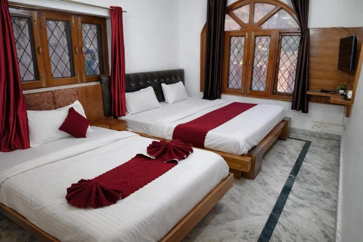 Ein Hotelzimmer mit zwei Betten, jeweils mit einem hölzernen Kopfteil und weißer Bettwäsche. An den Fenstern befinden sich rote Vorhänge und ein an der Wand befestigtes Fernsehgerät.