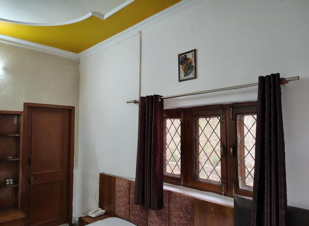 A room with a wooden door, a window with curtains, a small table with a telephone, and a wall-mounted picture.