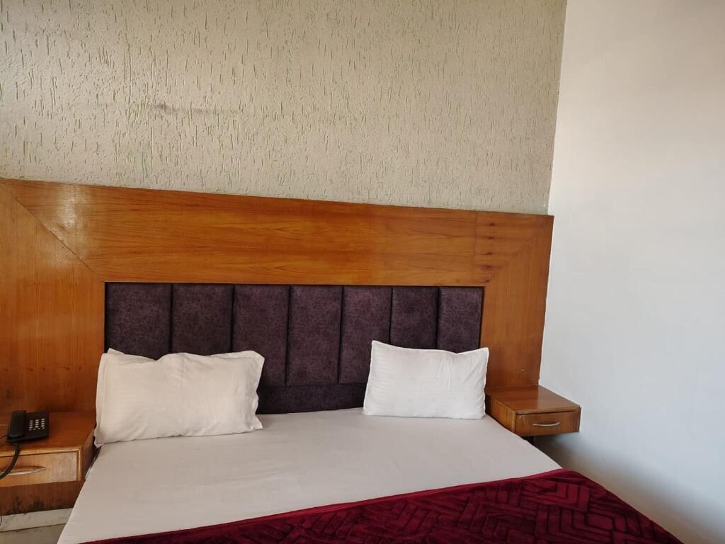 A hotel room with a wooden headboard, two white pillows, a red patterned bedspread, and a bedside table with a phone.