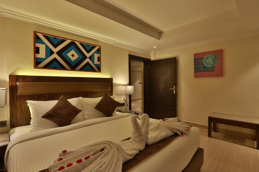 A hotel room with a large bed, a wooden headboard, a wall-mounted artwork, a bedside table with a lamp, and a door.